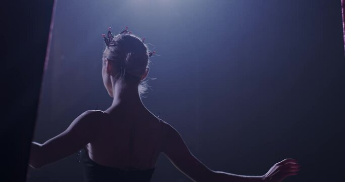 Rear on Caucasian graceful and famous ballerina in black dress and crown coming out on stage from curtains and bowing. Back view on female ballet dancer entering the stage to bow.