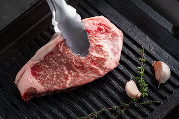 Raw Striploin steak is placed with tongs on a grill pan with garlic and thyme on a dark background, close up