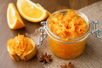 A jar of orange confiture, slices of orange fruit, pieces of French baguette with star anise on the sackcloth and wooden background