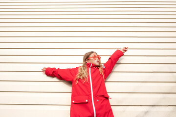 Girl with sunglasses and red jacket standing near white wall. Spending time outdoors. Child playing outside in autumn. Childhood memories. Going on journey, adventure in wilderness.