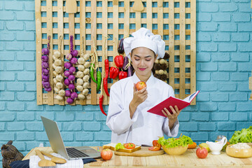 Young woman chef cooking in kitchen.An Asian woman is training to make a sandwich with a vegetable salad from the internet on a laptop in the kitchen with vegetables on the table.