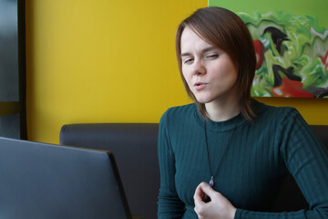 A girl with a pensive tense expression on her face sits, working at a laptop at a table in a cafe on a brown sofa against a yellow wall. Holds a jewelry on his chest with his hand.