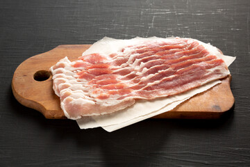 Uncooked Raw Bacon on a rustic wooden board on a black surface, side view. Close-up.