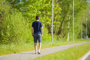 Fototapeta na wymiar One young man in shorts and t-shirt slowly walking on long sidewalk at town green park. Warm, sunny spring day. Spending time alone in nature. Peaceful atmosphere. Back view.