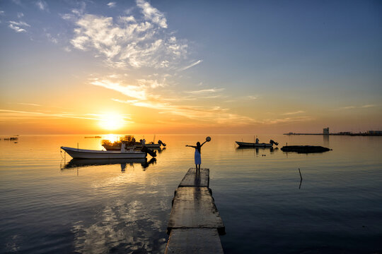 Silhouette happy woman holding hat open arms on to sky standing on pier on sea water sunrise colorful view with baots floating on water background, Bahrain.