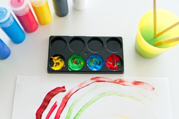 Paper with a child's drawing with several containers of acrylic paint and a plastic cup with water