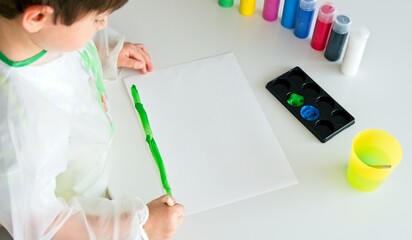 Boy painting with a brush and green paint a white paper