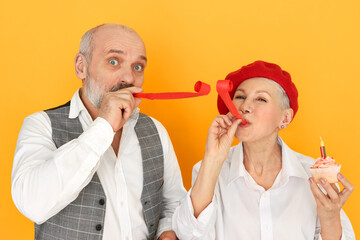 Successful romantic senior couple celebrating marriage anniversary. Studio shot of handsome elderly man and mature female in red bonnet blowing whistles, having fun, eating birthday cupcake