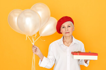 Portrait of sad unhappy mature woman in elegant clothes posing agaisnt yellow background with box of chocolate and helium balloons, giving birthday present, having upset depressed expression