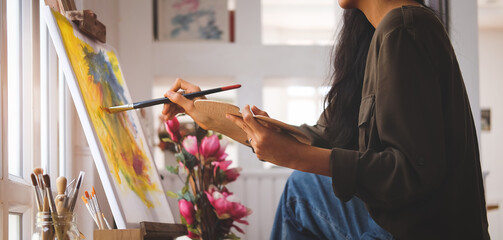Cropped shot view of Woman painter sitting on the floor in front of the canvas and drawing. Artist studio interior. Drawing supplies, oil paints, artist brushes, canvas, frame.