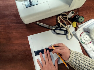 Seamstress draws a pattern with a pencil and a ruler on a white sheet of paper against the sewing supplies. Top view on a brown wooden table with female hands, a sewing machine, scissors, cloth, etc