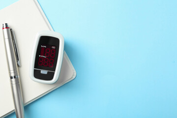 Modern fingertip pulse oximeter, pen and open notebook on light blue background, flat lay. Space for text