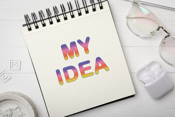 Notebook with words MY IDEA, glasses and earphones on white wooden table, flat lay