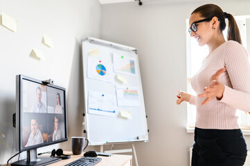 Webinar, online meeting, presentation. A young woman speaks to the online audience via video call, video connection. She stands in front of pc screen with online viewers, flip chart on the background