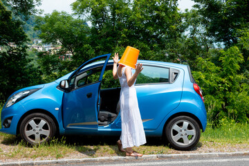 Woman with an orange bucket on her head gets out of the car because of a breakdown. She is...