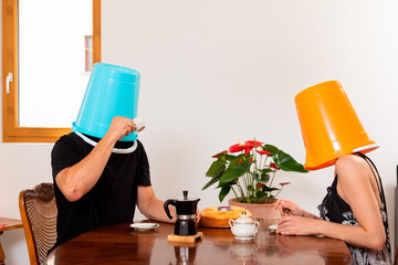 Young loving couple drinks coffee in the dining room with a bucket on their heads.