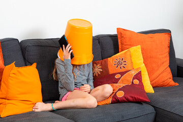 Little girl sitting on a sofa at home with a bucket on her head plays with the tablet