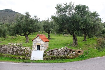 Fototapeta na wymiar A small country iconostasis, church-like small-scale replica for candle-lighting with olive grove in the background in Messinia region, southwestern Greece.