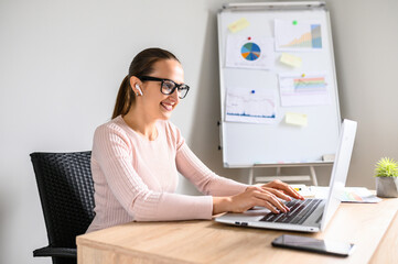 Happy young woman in smart casual works in the office, she typing on laptop keyboard and looks at screen with smile. Flip chart on the background