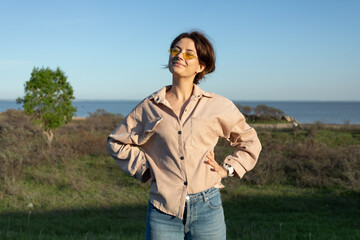 Posing woman in nature. Young woman in stylish clothes:  shirt and sunglasses  smilling   against the backdrop of a  sea, forest and field. Photos on summer vacation