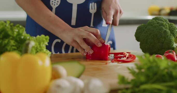 Close-up shooting. A woman is slicing a large pepper on the kitchen board. Salad preparation. There are a lot of vegetables around. 4K