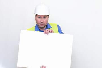 Close-up Engineer man wearing a helmet and reflector vest, standing and shows blank  billboard. White studio wall.
