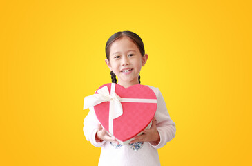 asian little child girl holding red heart gift box isolated on yellow background. Kid giving red heart gift box for you. Concept of love