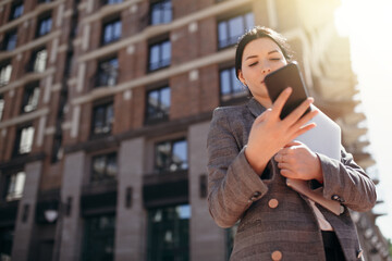 Fototapeta premium Bottom view of a businesswoman typing a message on her smartphone and holding a laptop in her hands