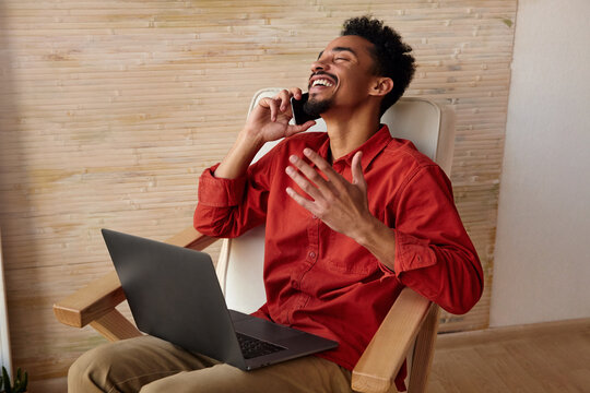 Overjoyed young pretty short haired bearded dark skinned man thowing back his head while laughing and raising emotionally hand, making phone call while sitting in chair