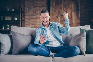 Photo of handsome guy stay home quarantine time hold telephone blogger freelancer good mood check likes followers legs crossed sit comfy sofa modern interior living room indoors