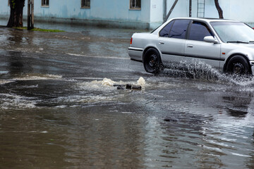 Вriving car on flooded road during flood caused by torrential rains. Cars float on water, flooding streets. Splash on car. Flooded city road with large puddle.  Flooding after heavy rains at city