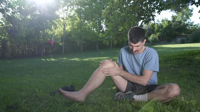 Male runner in pain on injury at the knee the leg on a park exercising or practicing for a marathon race, wearing sport clothes on sunny sunset sky. Spring time.