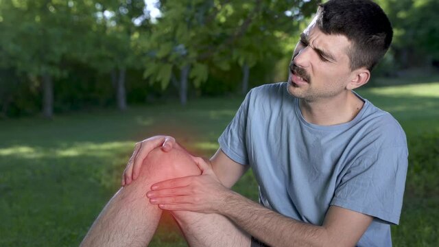 Male runner in pain on injury at the knee the leg on a park exercising or practicing for a marathon race, wearing sport clothes on sunny sunset sky. Spring time.