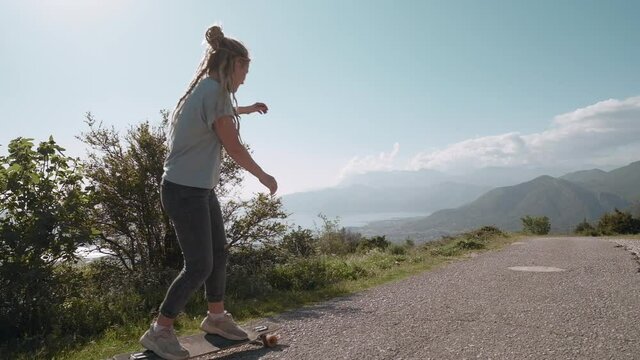 Back view traveler dreadlocks woman longboarding on mountain background riding skateboard cruising downhill on countryside road having fun enjoying relaxed summer vacation, Montenegro
