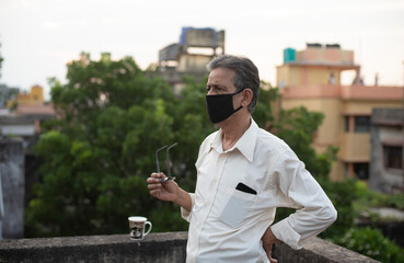 Portrait of an Indian old man with corona preventive mask, specs in hand and a coffee mug on rooftop during afternoon in home isolation.Indian lifestyle, disease and home quarantine.