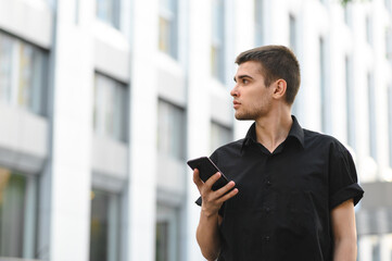 Attractive young office worker in a black shirt stands on a light urban background with a smartphone in his hand and looks away at a blank space. Portrait of stylish office worker