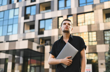 Handsome guy in casual clothes and with a laptop in his hand stands on the street, looking up with a serious face.Street portrait of a fashionable man holding a laptop, wearing a black shirt