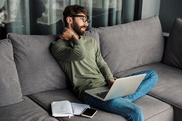 Image of focused bearded man working with laptop while sitting on sofa