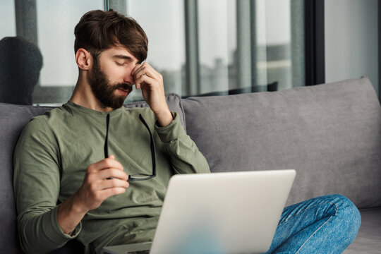 Image of tired bearded man holding eyeglasses and working with laptop