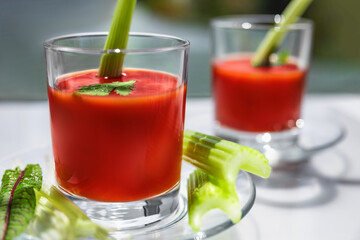 Two glasses of tomato juice with parsley and celery decorations, stand near the window, morning sunlight shines, shallow depth of field, selective focus. Natural drinks concept.
