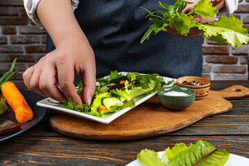 Woman's hands decorate and prepare a fresh green salad in a plate with spinach, arugula, carrot romance, on an old wooden table. Natural healthy food concept.