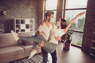 Photo of young couple guy hold arms excited lady quarantine together stay home hugging happy have fun just married moved new flat wear facial medical mask living room evening sunset indoors