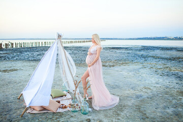 Fototapeta na wymiar Beautiful young pregnant woman with long blond hair in pink dress on the beach touching her belly with love and care on the background of sea. Blur sea background. Maternity, childbirth concept
