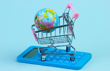 Global supermarket, shopping time. Supermarket trolley with globe and calculator on blue studio background.