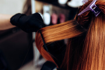 Hairdresser straightens redhead hair of woman with hair straightener tool in hair salon.
