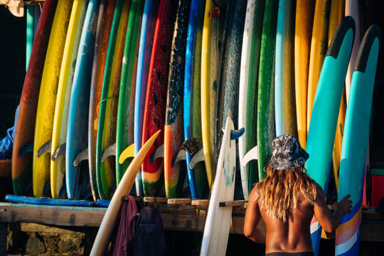 Set of different color surf boards in a stack by ocean with unrecognizable man choosing board. Bali.Indonesia. Surf boards on sandy beach for rent. Surf lessons on Weligama beach, Sri Lanka.