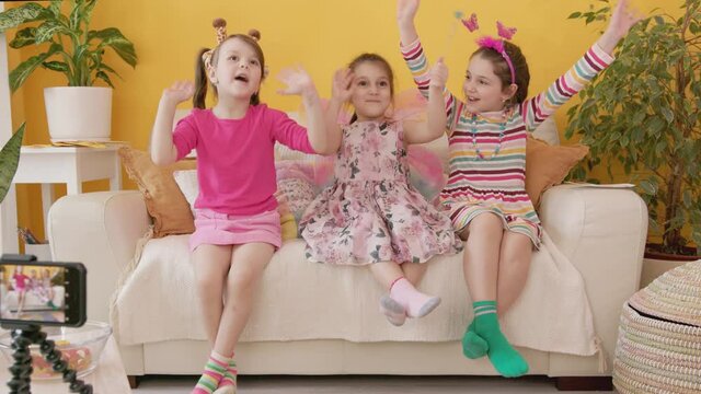 Shooting a Video Blog for Children with Smartphone. Production of Kid's Content. Three Cute Cheerful Little Caucasian Girl Talking and Laughing, Sitting on a Couch. Cozy Interior with Yellow Wall.