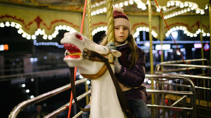 Fototapeta na wymiar Sad kid girl riding a carousel on a white horse at night