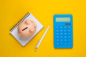 Piggy bank, notebook and calculator on yellow background. Minimalistic studio shot. Overhead view....