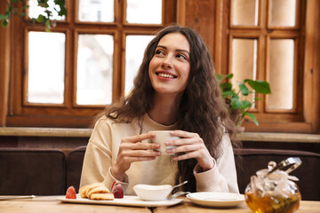 Image of happy woman smiling and drinking tea while sitting in cafe